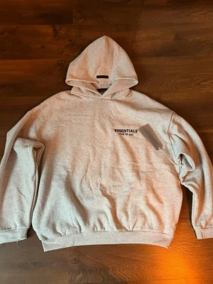 Grå Essentials Fear of God hoodie - Snygg grå hoodie från Essentials Fear of God med svart logga på bröstet och stor text på ryggen. Oversized passform, mjukt material och klassisk huva. Perfekt för en avslappnad streetwear-look.