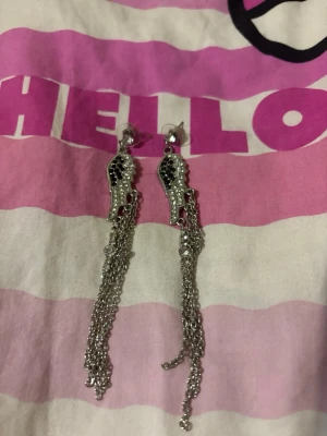 Silvriga örhängen med kedjor och stenar - Säljer ett par silvriga örhängen med hängande kedjor och gnistrande svarta och klara stenar. Örhängena har en unik form upptill och flera tunna kedjor som ger en cool och trendig look. Perfekta för dig som vill sticka ut lite extra.