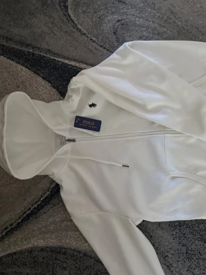 Vit hoodie från Polo Ralph Lauren - Snygg vit hoodie från Polo Ralph Lauren med dragkedja framtill och klassisk huva med dragsko. Märkets ikoniska logga är broderad på bröstet. Hoodien har en stilren look och är tillverkad i mjukt material för maximal komfort.