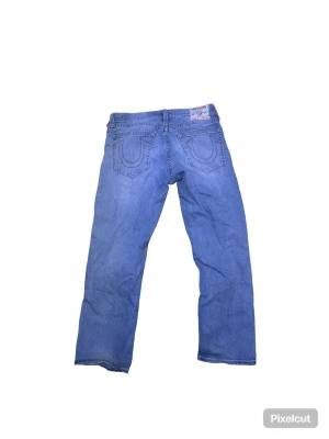 Blå raka jeans från True Religion - Säljer ett par klassiska blå jeans från True Religion med raka ben och ikoniska sömmar på bakfickorna. Jeansen har normal midja, fem fickor och är gjorda i mjukt denimtyg. Perfekta för en avslappnad och trendig look.