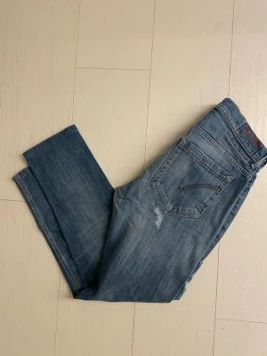 Dondups - Säljer ett par blå jeans med raka ben och diskreta slitningar på baksidan. Jeansen har klassiska bakfickor med dragkedjedetaljer och är tillverkade i denim. Perfekta för en avslappnad och trendig look.