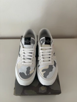 Valentino vita camo sneakers - Snygga sneakers från Valentino i vitt med grått och beige camomönster. Skorna har låga skaft, vita snören och svart logga på plösen. Tillverkade i en mix av läder och textil för en clean och modern look.