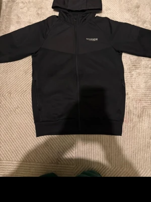 Svart hoodie från McKenzie - Snygg svart hoodie från McKenzie med dragkedja framtill och huva. Tröjan har diskret logga på bröstet och två fickor på sidorna. Perfekt för en avslappnad streetstyle-look.