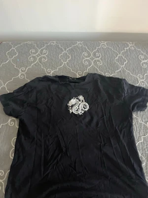 Svart t-shirt med drakbrodyr - Snygg svart t-shirt med en vit broderad drake på bröstet. T-shirten har korta ärmar och en klassisk passform. Perfekt för dig som vill ha en unik detalj i din outfit.