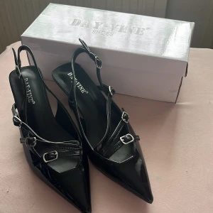 Svarta kitten heels från DAY-VINE - Snygga svarta pumps i lack från DAY-VINE med spetsig tå, låg klack och flera smala remmar med silvriga spännen. Skorna har öppen häl och elegant design som ger en trendig vibe. Perfekta för dig som vill sticka ut med stilrena detaljer. Köpta från Raglady By Tara.