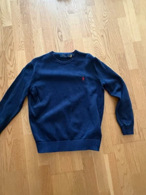 Mörkblå stickad tröja Polo Ralph Lauren - Mörkblå stickad tröja från Polo Ralph Lauren med rund halsringning och ribbade muddar. Tröjan har den klassiska röda logotypen broderad på bröstet. Perfekt för dig som gillar stilrena och tidlösa plagg.