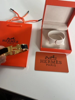 Svart och guldarmband från Hermès - OBS byggmiss och går inte att stänga ordentligt därav priset. Snyggt svart armband från Hermès med guldfärgad metall och den ikoniska H-detaljen framtill. Kommer i originalask och med Hermès påse. Perfekt accessoar för dig som gillar lyxiga och stilrena smycken.