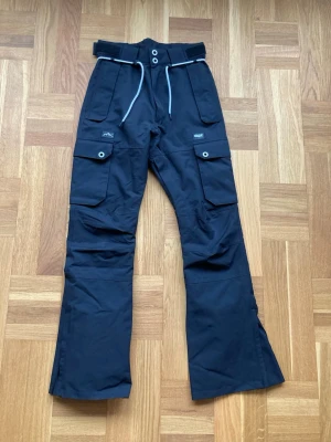 Svarta snowboardbyxor från Dope - Svarta täckbyxor från Dope med flera stora fickor, justerbar midja med snörning och tryckknappar. Vattentäta och ventilerande, tillverkade i delvis växtbaserat material. Perfekta för vinterns äventyr i backen eller på fjället.