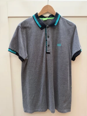 Grå pikétröja från BOSS - Snygg grå pikétröja från BOSS med turkosa och svarta detaljer på krage och ärmslut. Tröjan har korta ärmar, knappar framtill och BOSS-logga broderad på bröstet. Tillverkad i mjuk bomull för en bekväm känsla. Knappt använd. 