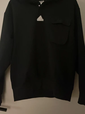 Svart fodrad hoodie från Adidas med ficka - Snygg svart fodrad hoodie från Adidas med klassisk logga på bröstet och en praktisk bröstficka. Hoodien har huva och långa ärmar, perfekt för en avslappnad streetwear-look. Tillverkad i mjukt material som känns skönt mot huden.
