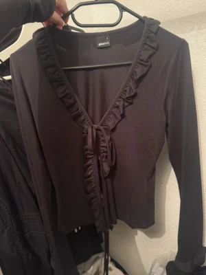 Svart volangtopp från Gina Tricot - Supersnygg svart topp från Gina Tricot med långa ärmar, v-ringning och volangkant längs ringningen. Toppen har knyt framtill, vilket ger en trendig look. Perfekt att styla med höga jeans eller kjol.