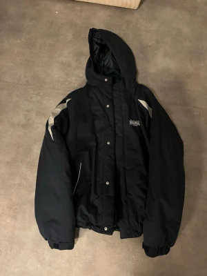 Svart vindjacka från Lonsdale - Snygg svart vindjacka från Lonsdale med huva och silverfärgade detaljer på axlar och rygg. Jackan har tryckknappar framtill, resår i ärmslut och nederkant samt stor logga på ryggen. Perfekt för blåsiga dagar och streetwear-stil.