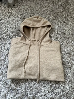 Beige merinoull hoodie med snören och skönt material - Snygg beige merinoull hoodie med huva och dragsko. Tillverkad i mjukt stickat material som känns skönt mot huden. Hoodien har ribbade muddar vid ärmslut och nederkant för en clean look. Perfekt till jeans eller joggers för en chill stil.