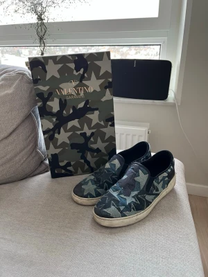 Valentino Garavani camo slip-on sneakers - Snygga slip-on sneakers från Valentino Garavani med ett unikt kamouflagemönster i blå, grå och svarta toner. Skorna har vita sulor och svarta detaljer vid hälen. Tillverkade i textil och läder för en cool och modern look.