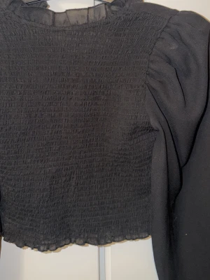 Svart smockad blus med volangkrage - Trendig svart blus med smockad framsida och volangdetalj vid kragen. Blusen har puffiga långärmade ärmar i ett tunt, lätt material som ger en luftig känsla. Perfekt att styla med både jeans och kjol för en cool look.