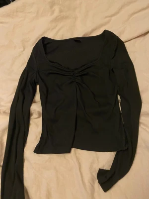 Svart croppad långärmad topp - Trendig svart croppad topp med lång ärm och snygg rynkning vid bysten. Toppen har en bred, fyrkantig ringning och är tillverkad i ett ribbat material som ger en skön och stretchig passform. Perfekt att styla med höga jeans eller kjol.