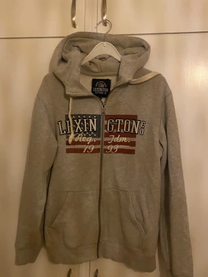 Grå Lexington zip-hoodie med tryck - Säljer en grå zip-hoodie från Lexington med stor huva och vita dragsnören. Framsidan har ett stort tryck i blått, vitt och rött med texten 'Lexington Clothing Co.' och amerikanskt flaggmotiv. Hoodien har två fickor fram och är gjord i mjuk bomullsblandning. Storlek M men sitter mer som S. Jag bär S och sitter perfekt på mig.