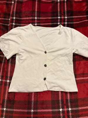 Vit v-ringad topp med knappar från Lager 157 - Stilren vit topp med v-ringning och tre knappar framtill. Kortärmad och i mjukt bomullsmaterial, perfekt för en enkel och clean look. Passar till både jeans och kjol för en avslappnad stil.