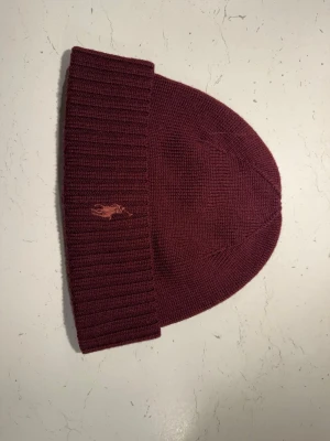Vinröd mössa från Polo Ralph Lauren - Säljer en vinröd ribbstickad mössa från Polo Ralph Lauren med klassisk broderad logga framtill. Mössan är i mjukt stickat material och har en bred uppvikt kant för extra stilpoäng. Perfekt accessoar för kalla dagar.