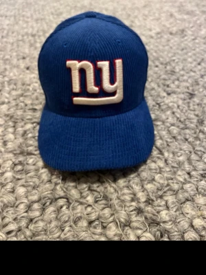 Blå New Era Giants keps i manchester - Snygg blå keps från New Era med broderad 'ny' logga framtill och Giants-tema. Kepsen är gjord i manchestertyg som ger en cool retrovibe. Perfekt för dig som gillar sportiga accessoarer och vill sticka ut med en unik look.