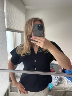 Svart blus med knappar och v-ringning - Stilren svart blus med v-ringning och två dekorativa knappar framtill. Blusen har korta, vida ärmar och en lätt figursydd passform. Perfekt för en trendig och enkel look. Materialet ser ut att vara ett mjukt tyg, troligen polyester eller bomullsblandning.