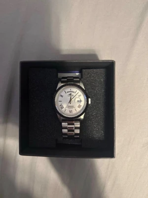 Silverfärgad Lotus klocka med vit urtavla - Stilren klocka från Lotus med silverfärgad länk och vit urtavla. Klockan har romerska siffror, datumvisning och klassisk rund form. Perfekt accessoar för dig som gillar en clean och tidlös look.