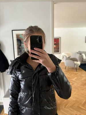 Svart pufferjacka från Peak Performance - Snygg svart pufferjacka från Calvin Klein med glansig finish och hög krage. Jackan har en klassisk pufferdesign med breda kanaler och dragkedja framtill. Perfekt för kalla dagar och riktigt trendig look.