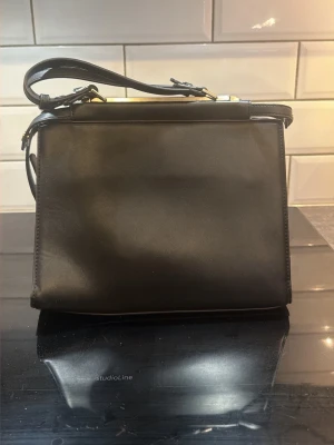 Svart klassisk handväska i faux-skinn - Snygg svart handväska i slätt skinn med justerbar axelrem och guldfärgade metalldetaljer. Väskan har en rak, boxig form och en praktisk ytterficka med dragkedja på baksidan. 3 fack på insidan där mittenfacket har två mindre öppna fickor och en större ficka med dragkedja. Perfekt för dig som gillar stilrena accessoarer.