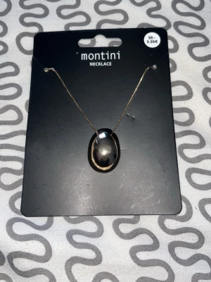 Guldfärgat halsband med svart sten - Snyggt halsband från Montini med en tunn guldfärgad kedja och en oval, svart sten som hänge. Perfekt accessoar för att lyfta din outfit med en stilren och modern touch.