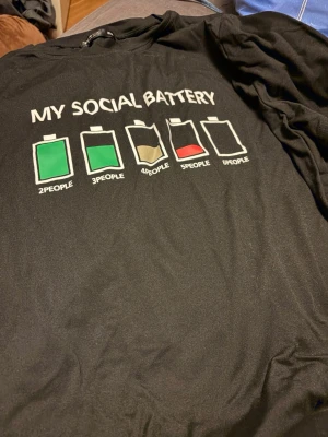Svart t-shirt med social battery-tryck - Rolig svart t-shirt med trycket 'MY SOCIAL BATTERY' och batteriikoner i grönt, beige och rött. Perfekt för dig som gillar humoristiska plagg och vill visa din personlighet. Klassisk passform och rund hals.