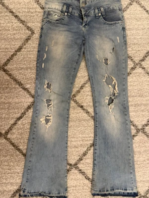 Ljusblå slitna jeans från Realist - Ljusblå jeans med slitningar och hål på benen från Realist. Modellen har bootcut och dubbla knappar i midjan. Snyggt tvättad denim med trendiga detaljer och broderad logga på insidan av linningen. Jeansen håller dock på att bli ett o samma hål på högra benet som man ser men går o fixa super enkelt med en säkerhetsnål, eller sy (inget som syns). Tryck inte Köp innan ni kontaktat mig Pris diskuteras!