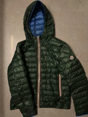 Moncler Jacka  - Tja! Säljer denna stilrena moncler jacka med färg grön och blå. Jackan är i storlek 3, vilket motsvarar cirka m. Skicket är acceptabelt då den har slitna ställen lite runt om kring. Insidan har fläckar men ingen som syns vid användning. Jackan är köpt hos Sicurofashion som har äkthetserifiering vid försäljning. Priset är diskuterbart vid en snabb affär😀😀Vid frågor kontakta mig!🙌🙌🤩
