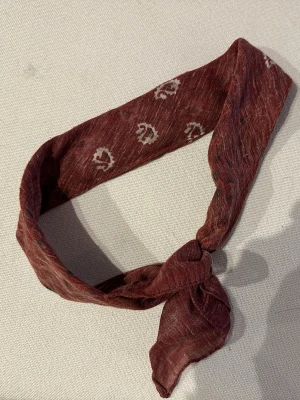 Vinröd tunn bandana med mönster - Säljer en vinröd, tunn sjal i mjukt tyg med vita abstrakta mönster. Sjalen är smal och lätt att knyta runt halsen eller i håret för en cool och avslappnad look. Perfekt accessoar för att lyfta din outfit.