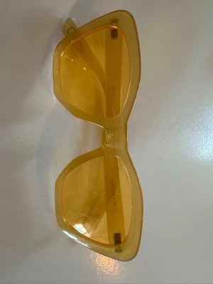 Gula solglasögon från Mango - Snygga solglasögon från Mango i en cool cat eye-modell med transparent gul plastbåge och matchande gula glas. Perfekta för dig som vill sticka ut och addera färg till din look.