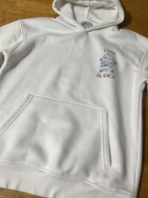 Vit hoodie Casa Blanca tryck - Vit hoodie från Casa Blanca med liten färgglad logga och text på bröstet. Klassisk känguruficka framtill och huva. Perfekt för en chill och stilren look. Mjuk bomullskänsla och relaxed fit.