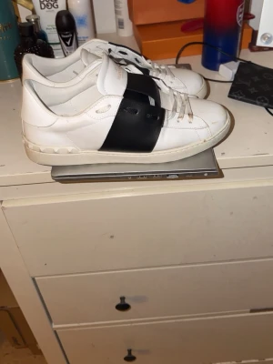 Valentino Garavani vita sneakers - Säljer ett par vita Valentino Garavani sneakers med svart band över mitten. Skorna har klassisk låg profil, vita snören och detaljerade nitar på hälen. Tillverkade i läder för en lyxig känsla och stilren look. Allt orginal kmr med är som nya. Tvättar innan frakt hör av dig för mer info.