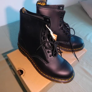 Svarta Dr. Martens boots stl 46 - Klassiska svarta Dr. Martens boots i skinn med ikonisk gul söm runt sulan och genomskinlig, räfflad sula. Högt skaft, snörning framtill och dragflik med logga bak. Perfekta för dig som vill ha en tidlös och edgy stil.