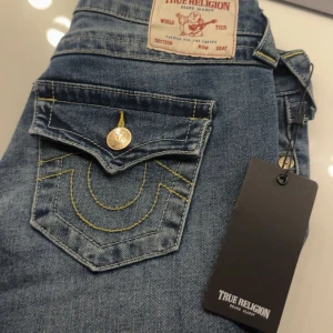 Blå jeans från True Religion - Snygga blå jeans från True Religion med ikonisk bakficka och gul söm. Jeansen har en klassisk femficksdesign och en knappdetalj på bakfickan. Varsamt använd, passar om man är petite, längden har lagts upp professionellt 