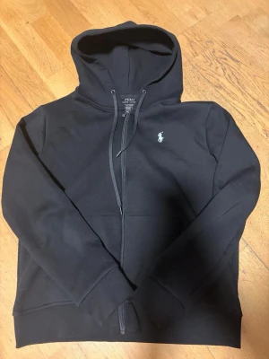 Svart hoodie från Polo Ralph Lauren - Snygg svart hoodie från Polo Ralph Lauren med dragkedja framtill och klassisk vit logga på bröstet. Hoodien har justerbar huva med snören och två fickor framtill. Tillverkad i mjukt bomullsmaterial som känns skönt mot huden.