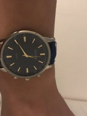 Blå klocka från Geneva med silverdetaljer - Stilren klocka från Geneva med mörkblå urtavla och matchande blått armband. Klockan har silverfärgat boett och spänne samt guldiga markeringar på urtavlan. Perfekt accessoar för dig som gillar klassisk och enkel design.