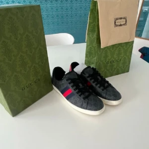 Gucci sneakers med monogram och röd detalj - Snygga Gucci sneakers i mörkgrått med klassiskt GG-monogram över hela skon. Skorna har svarta snören, vit sula och den ikoniska röd-blå randen på sidan samt röd detalj på hälen. Perfekta för dig som vill ha en lyxig och trendig look.