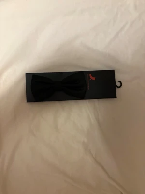 Svart fluga i klassisk design - Snygg svart fluga i klassisk form, perfekt för att lyfta din outfit till nästa nivå. Flugan är förknuten och sitter på ett justerbart band. Passar till skjorta och kavaj när du vill ha en stilren accessoar.