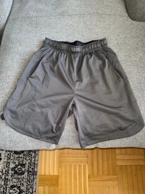 Grå träningsshorts från Nike - Snygga grå träningsshorts från Nike med elastisk midja och diskret logga på benet. Shortsen har en loose passform och är tillverkade i ett lätt och snabbtorkande syntetmaterial, perfekta för aktiva dagar.