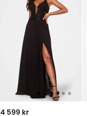 Svart långklänning med slits - Elegant svart långklänning med djup v-ringning och glittrig överdel. Klänningen har tunna axelband och en hög slits framtill som ger en snygg och modern look. Perfekt för bal eller festliga tillfällen.