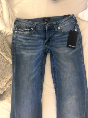 Blå bootcut jeans från True Religion - Säljer ett par snygga blå bootcut jeans från True Religion i modell Joey. Jeansen har klassisk femficksmodell, låg midja och slitningar för en cool vintage-look. Materialet är mjukt denim med lätt stretch för skön passform. 
