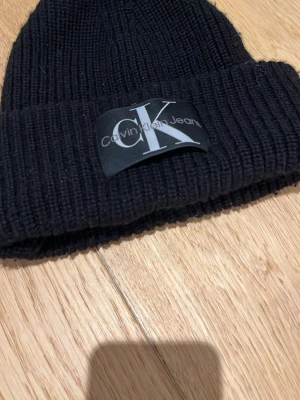 Svart ribbstickad mössa Calvin Klein - Säljer en svart ribbstickad mössa från Calvin Klein Jeans med stor CK-logga framtill. Klassisk uppvikt kant och mjukt material som håller dig varm. Perfekt accessoar för dig som gillar stilrena och tidlösa detaljer.