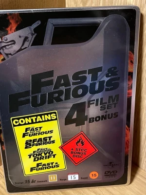 Fast & Furious 4-film DVD box - Säljer en Fast & Furious 4-film DVD box med bonusdisc i metallbox formad som en bensindunk. Innehåller de fyra första filmerna och en exklusiv STCC bonusdisc. Perfekt för dig som gillar action och bilar. Omslaget har svart och röd design med filmens logga.