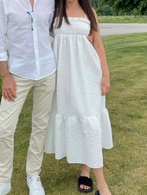 Vit långklänning med volang - Säljer en vit långklänning med smock upptill och tunna axelband. Klänningen har en volang nedtill och ett diskret strukturerat tyg som ger en somrig känsla. Perfekt för varma dagar och midsommarfirande.