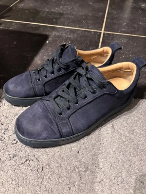 Mörkblå Louboutin skor - Snygga mörkblå sneakers i mocka med svarta skosnören och kontrasterande röd yttersula. Insidan är beige och skon har en klassisk låg profil med rund tå. Perfekta för dig som gillar stilrena detaljer och vill sticka ut med färg på sulan. Pris går att diskutera 