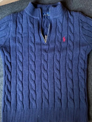 Mörkblå kabelstickad tröja Polo Ralph Lauren - Kabelstickad mörkblå tröja från Polo Ralph Lauren med halv dragkedja och hög krage. Tröjan har ett rött broderat logotyp på bröstet och ribbade muddar. Klassisk och tidlös design i mjukt material.